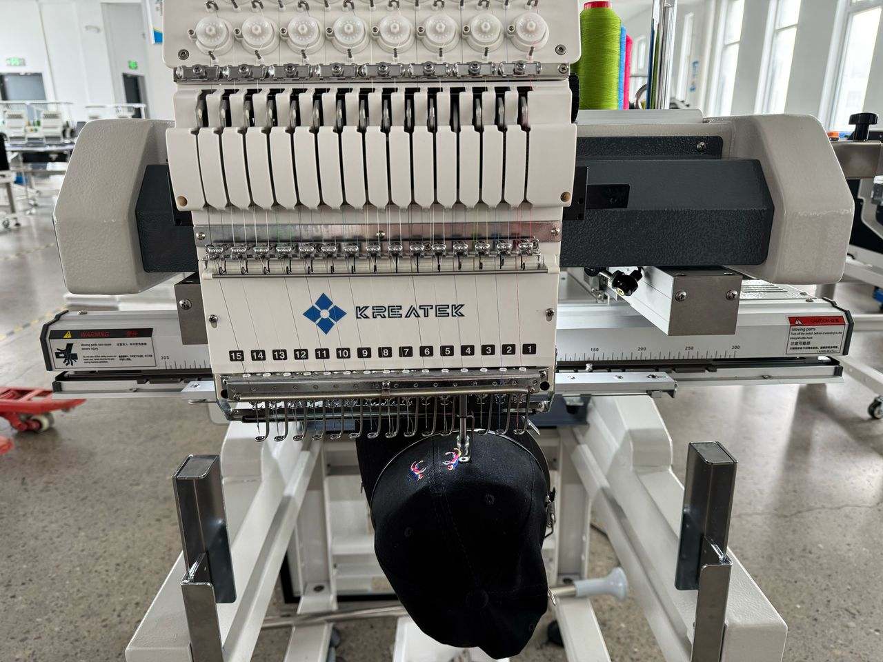 Kreatek BL-1501 industrial embroidery machine with 15-needle high-speed head stitching a black cap in a factory setting