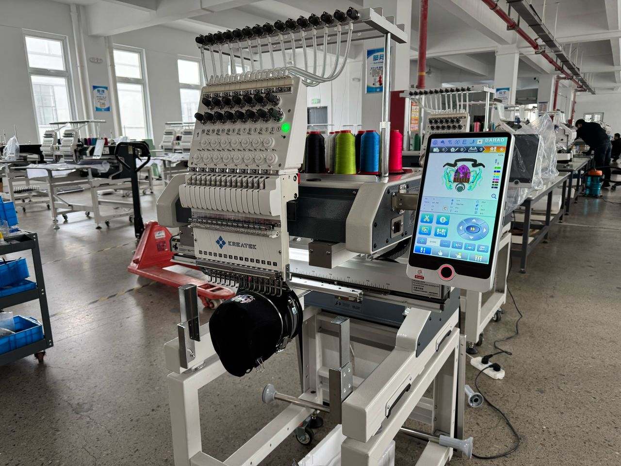 Kreatek III BL-1501 industrial embroidery machine with 12/15 needles, high-speed single head, digital touchscreen, and cap embroidery setup in a factory setting.