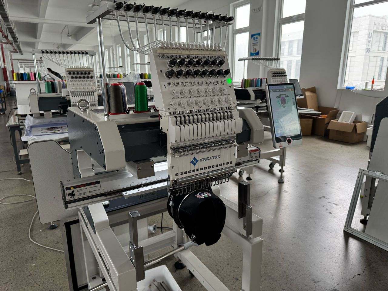 Kreatek III BL-1501 industrial embroidery machine with 15 needles, high-speed cap embroidery, digital screen, and Dahao control system in a textile workshop.