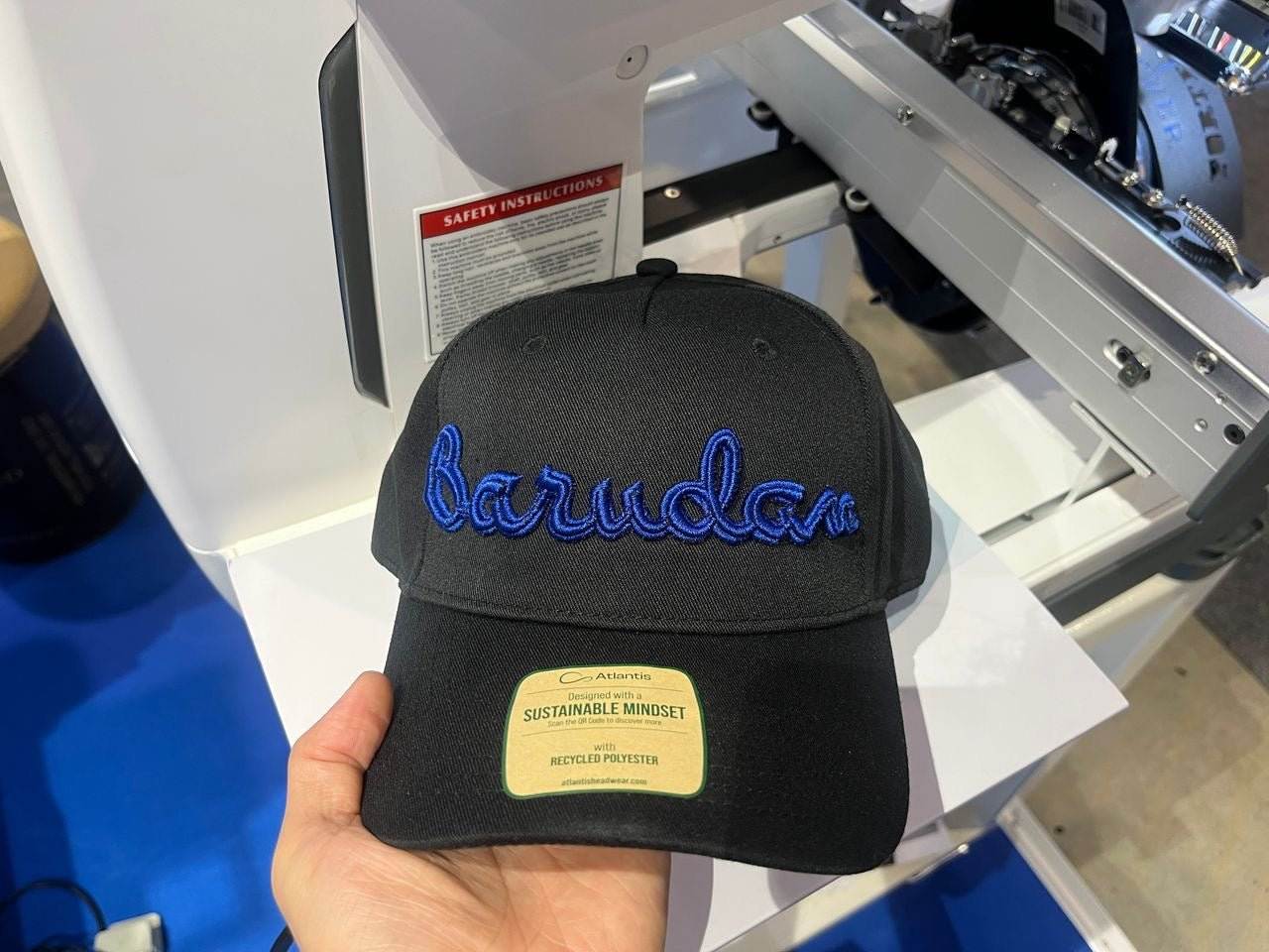Svart caps med blått brodert "Barudan"-tekst foran en industri broderimaskin som setter en ny standard innen profesjonell brodering. ✅